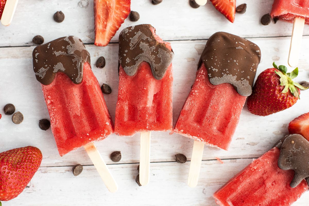 Four strawberry popsicles partially dipped in chocolate are arranged on a white surface, surrounded by fresh strawberries and chocolate chips.