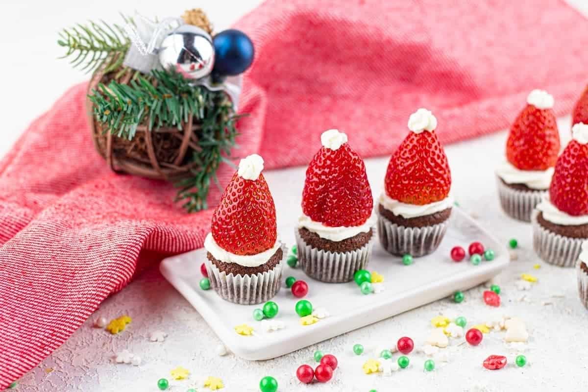 Mini chocolate cupcakes topped with white frosting and strawberries shaped like Santa hats, displayed on a white tray with festive decorations in the background.