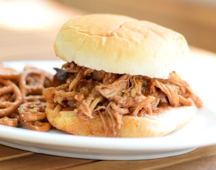 A pulled pork sandwich on a white bun served on a white plate with a side of pretzels.