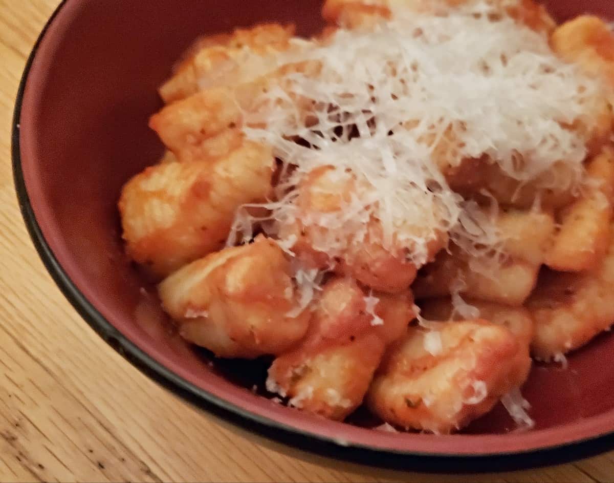 A bowl of gnocchi topped with tomato sauce and grated cheese, served in a red bowl on a wooden table.