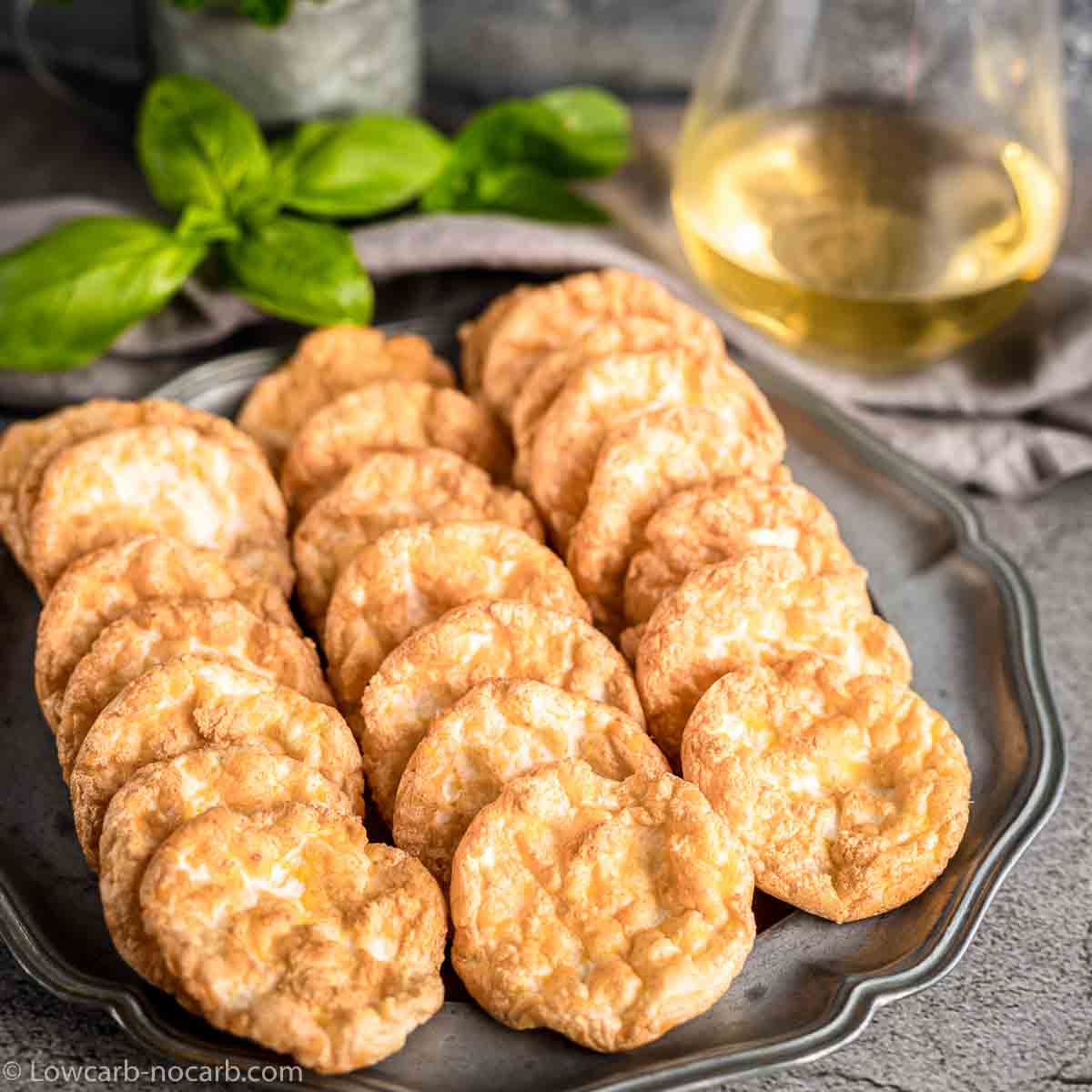 A metal tray holding rows of round, golden-brown cheese crisps, with a glass of white wine and green basil leaves in the background.