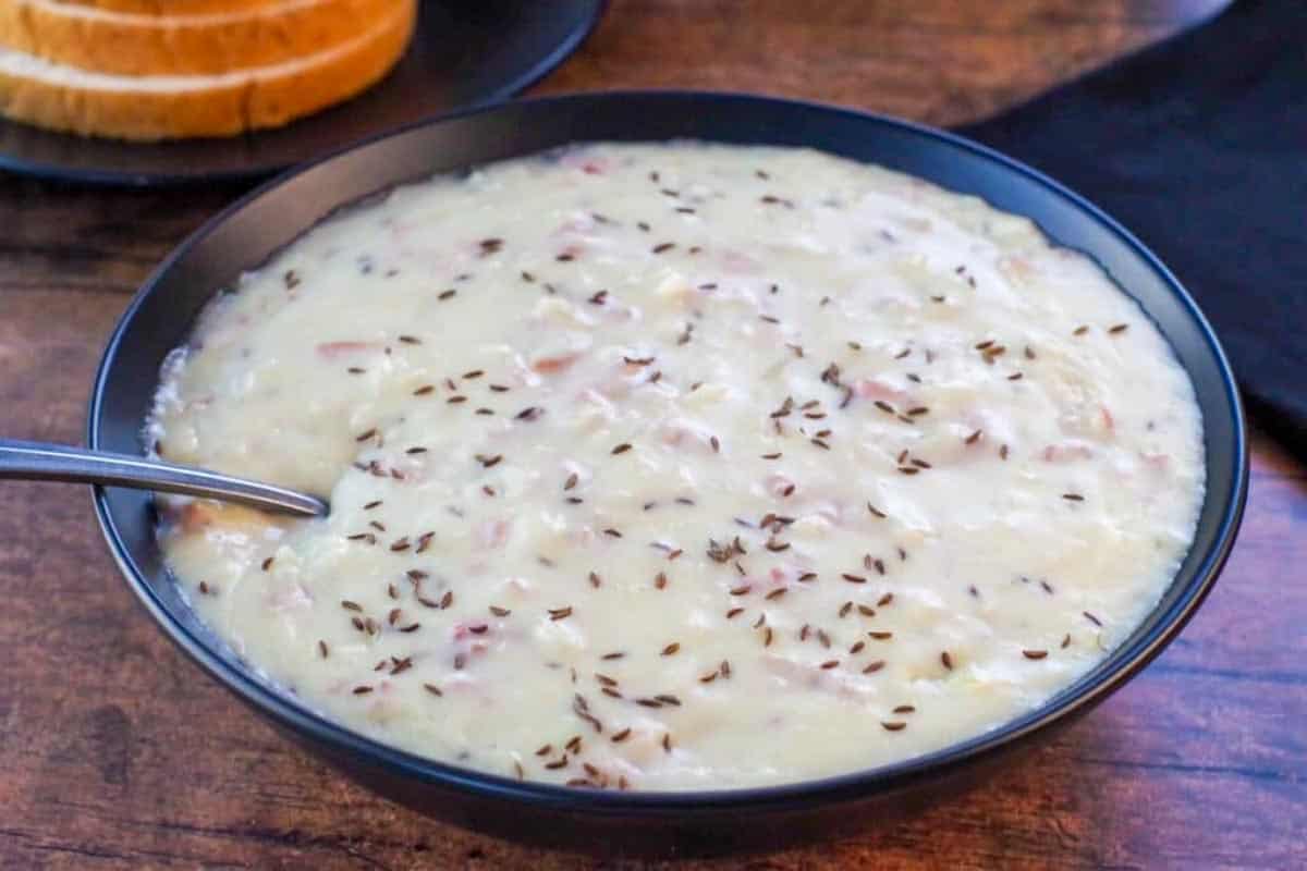 A black bowl filled with creamy soup topped with caraway seeds, with a spoon inside. Slices of bread are visible on a plate in the background.