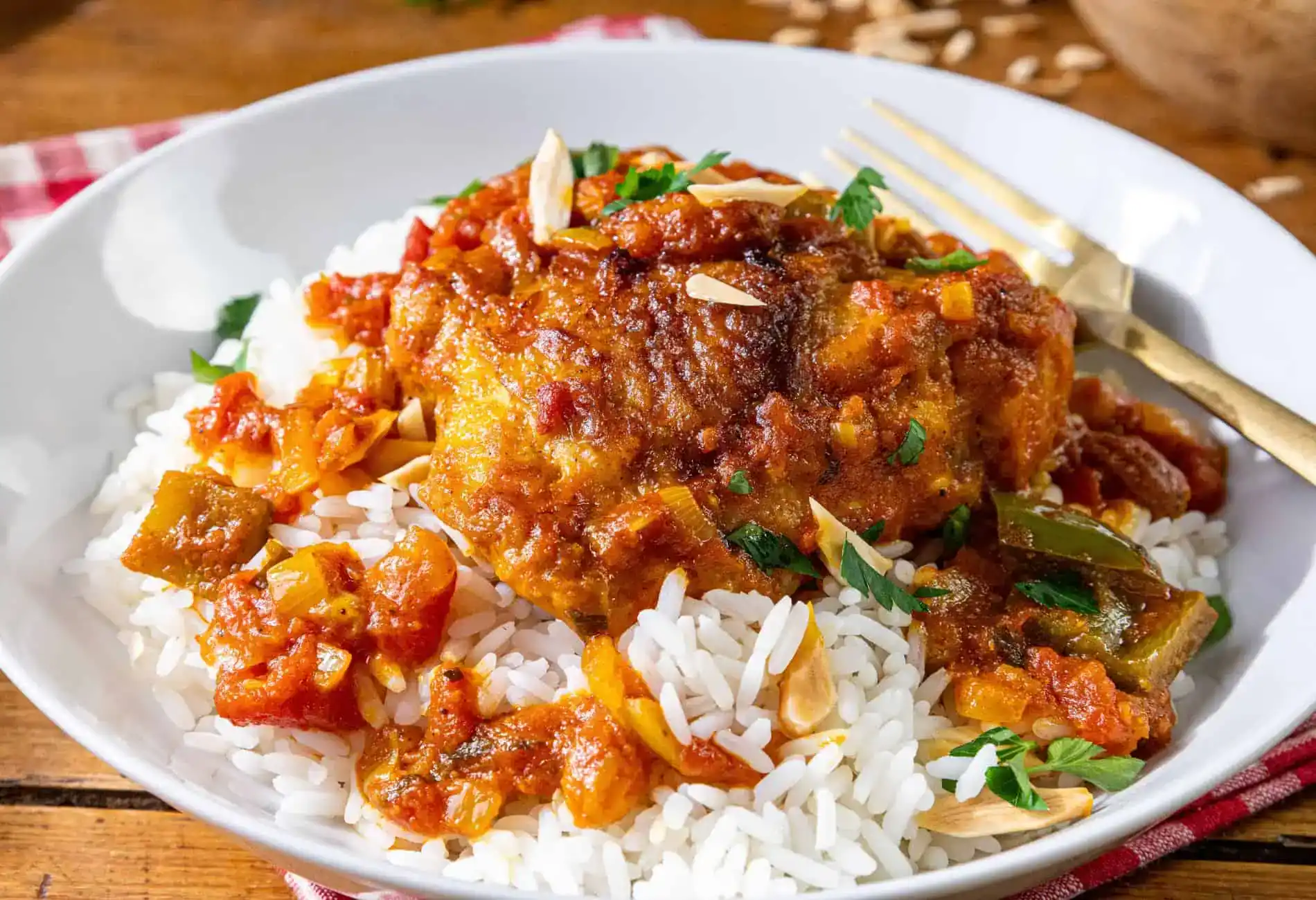 A plate of white rice topped with a piece of braised chicken in tomato-based sauce, garnished with chopped herbs and sliced almonds, with a gold fork on the side.