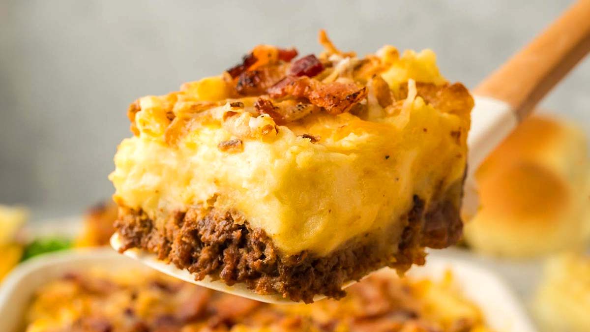 A close-up of a slice of casserole with a ground meat base, topped with mashed potatoes, melted cheese, and bacon pieces on a spatula.