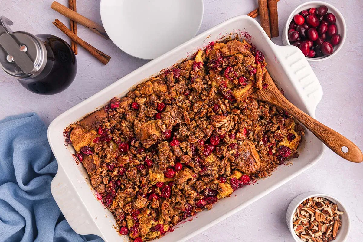 A white baking dish filled with cranberry pecan bread pudding, with a wooden spoon, surrounded by cinnamon sticks, maple syrup, cranberries, and chopped pecans.