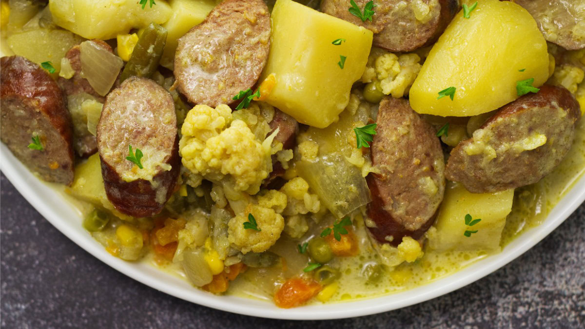 A close-up of a plate containing sliced sausage, potatoes, cauliflower, peas, carrots, and corn in a creamy sauce, garnished with chopped parsley.