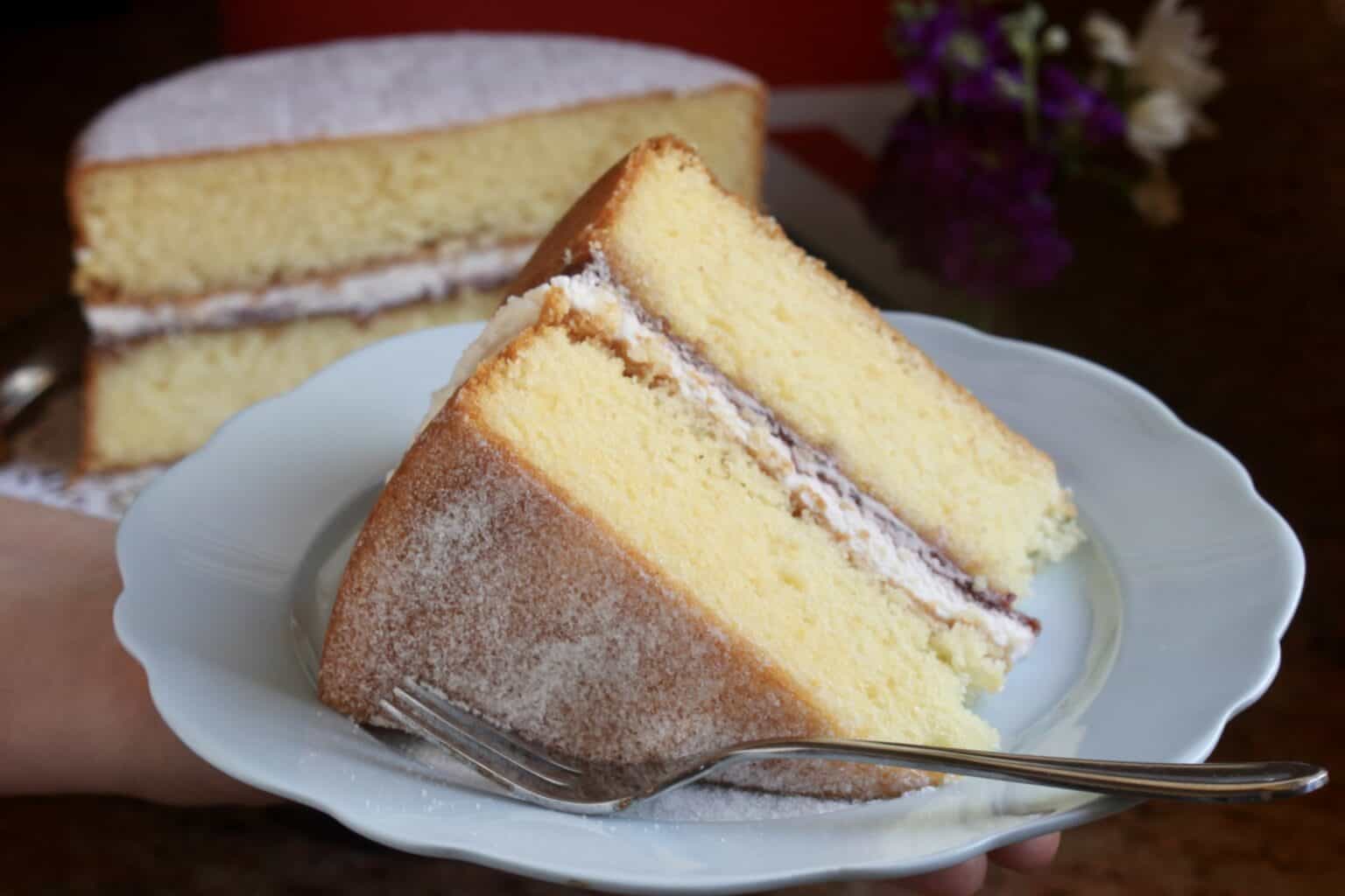 A slice of layered sponge cake with cream filling and powdered sugar on top, served on a white plate with a fork; the rest of the cake is in the background.