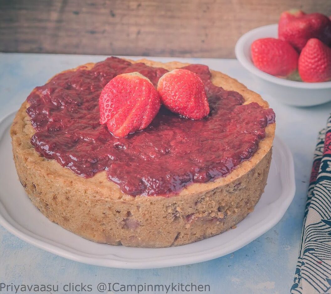 A round cake topped with strawberry jam and fresh strawberries sits on a white plate; a bowl of strawberries is in the background.