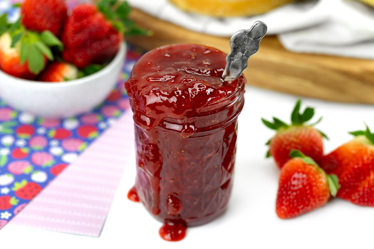 A jar of strawberry jam with a spoon inside, jam overflowing the rim, surrounded by fresh strawberries and a bowl of strawberries in the background.