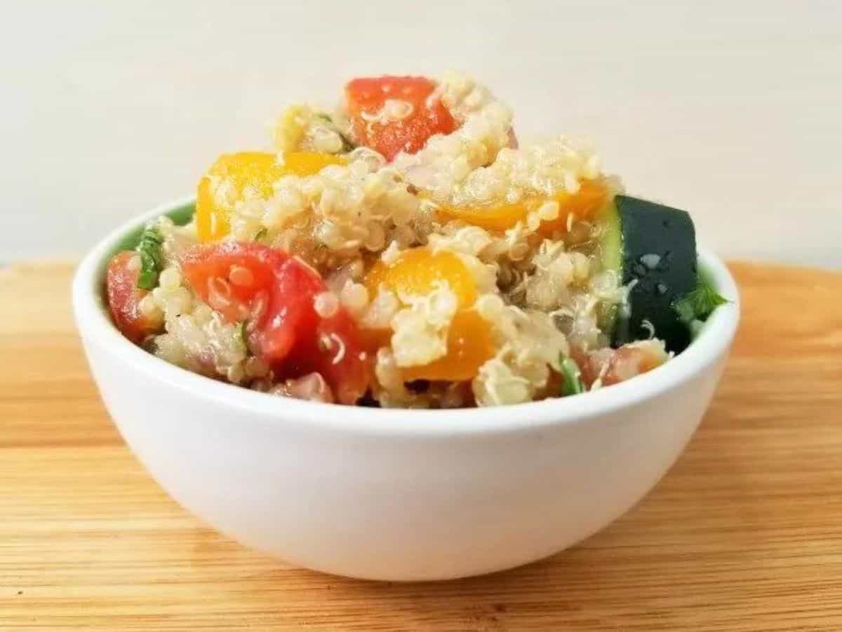 A white bowl filled with quinoa salad containing chopped yellow and red tomatoes and green vegetables, placed on a wooden surface.