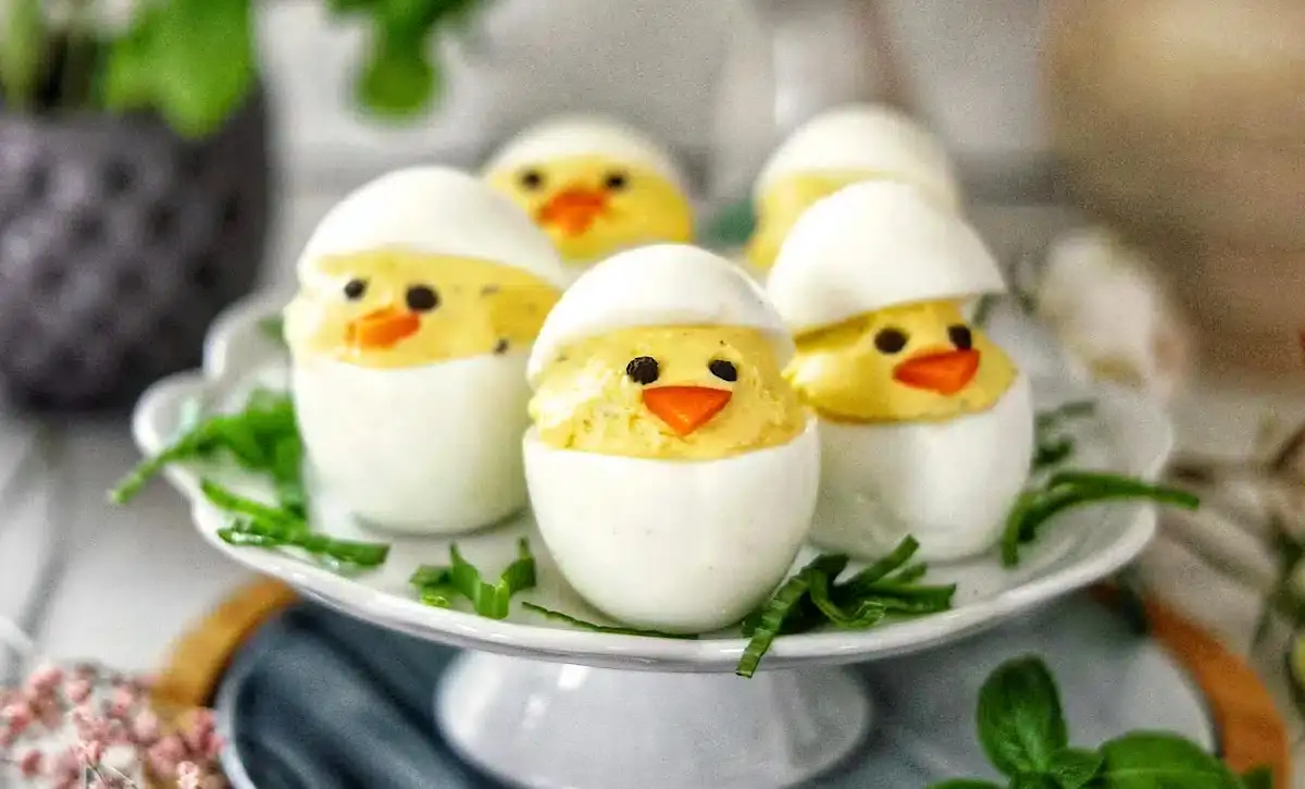 Deviled eggs arranged to look like chicks, with egg white halves as shells, yolk filling as faces, and small pieces of vegetables for eyes and beaks, displayed on a white plate.