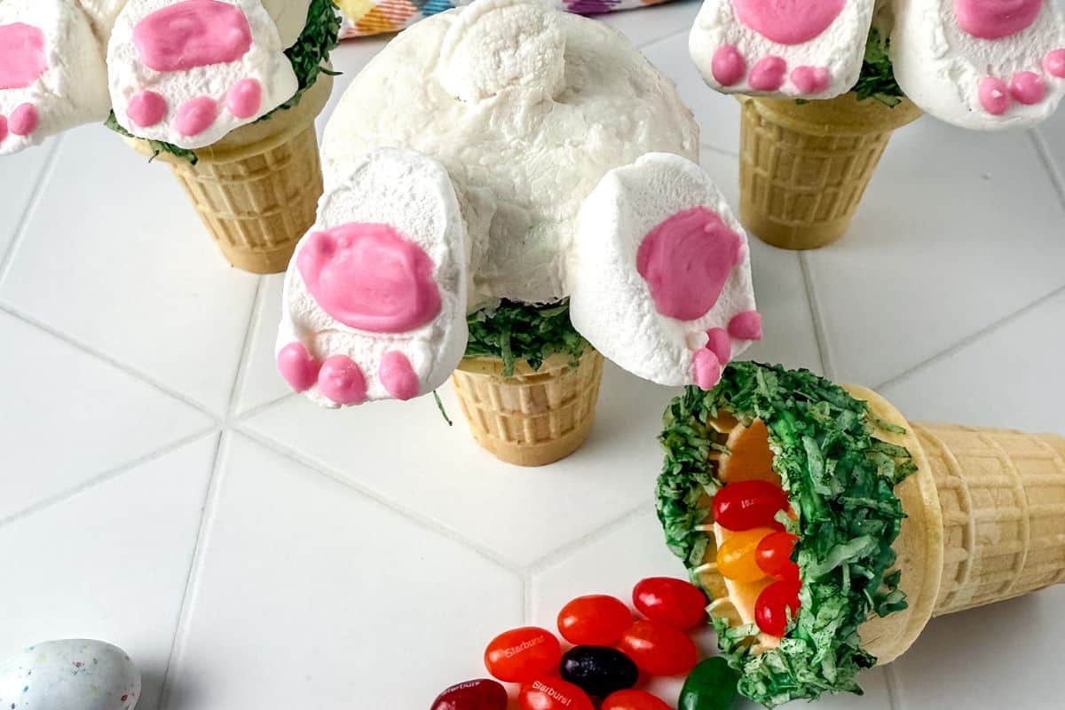 Ice cream cones topped with marshmallows decorated to look like bunny feet and a bunny tail, with colored jelly beans spilling from one cone.