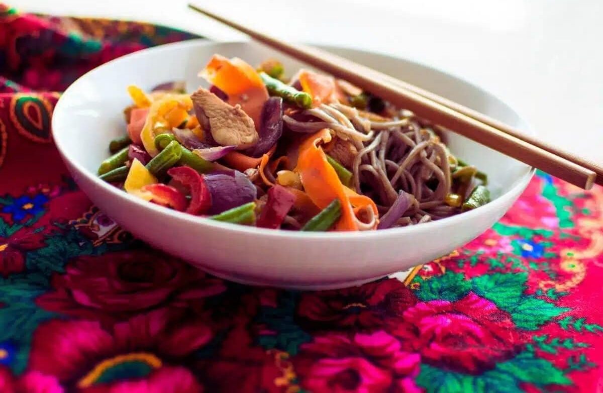 A bowl of soba noodles with sliced vegetables and meat, served with chopsticks on a colorful, floral-patterned cloth.