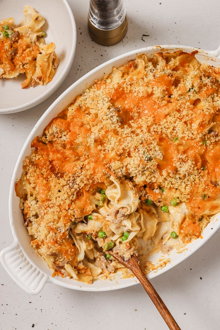 Baked noodle casserole topped with breadcrumbs and cheese in a white dish, with a serving spoon and a portion on a plate nearby.