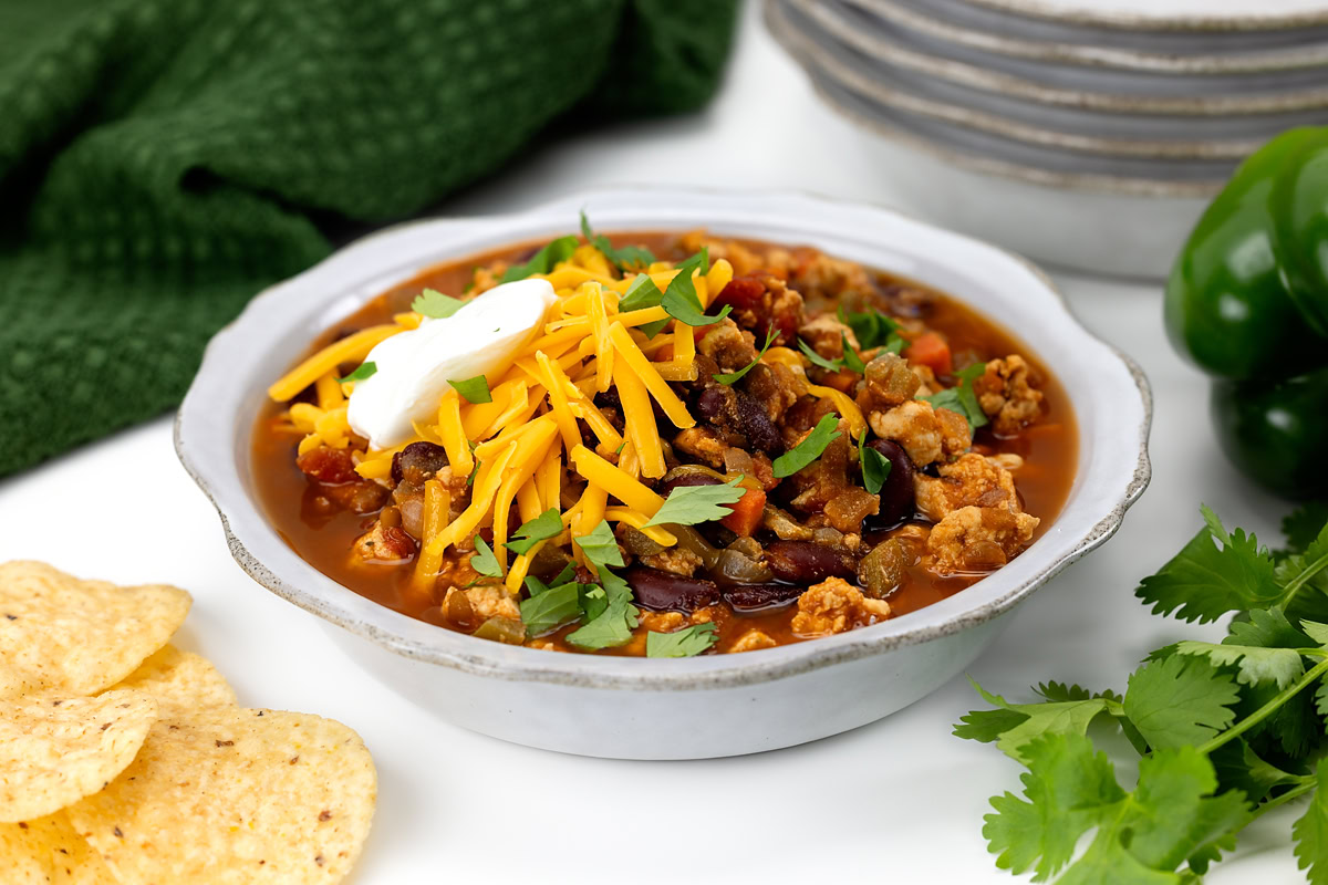 Bowl of chili topped with shredded cheddar cheese, sour cream, and chopped parsley, surrounded by tortilla chips and fresh cilantro.