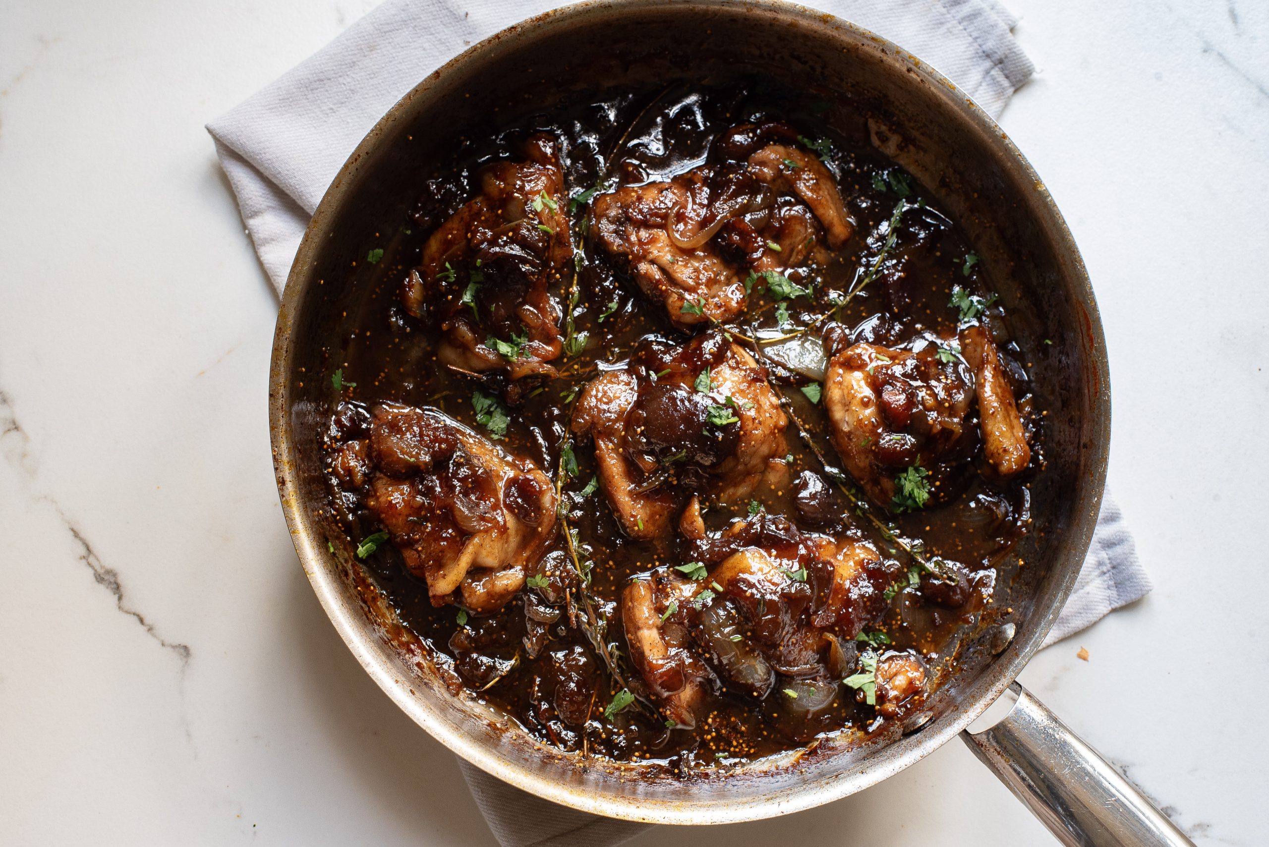 A skillet filled with cooked chicken thighs in a dark, glossy sauce sits on a white napkin on a light countertop.