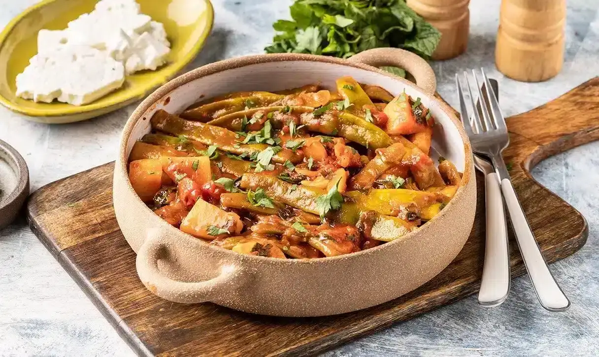 A bowl of stewed green beans, potatoes, and tomatoes garnished with herbs, placed on a wooden board with cutlery, herbs, and feta cheese on the side.