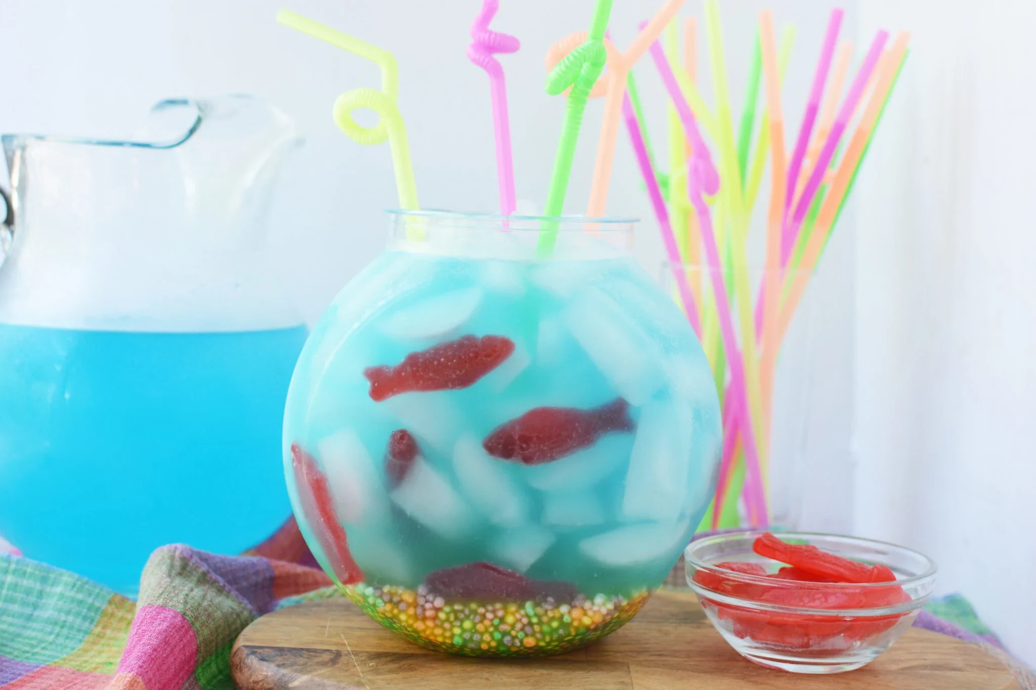 A round glass bowl filled with blue drink, ice, gummy fish candies, and colorful candy at the bottom. Several neon straws are in the bowl. A pitcher and extra straws are in the background.