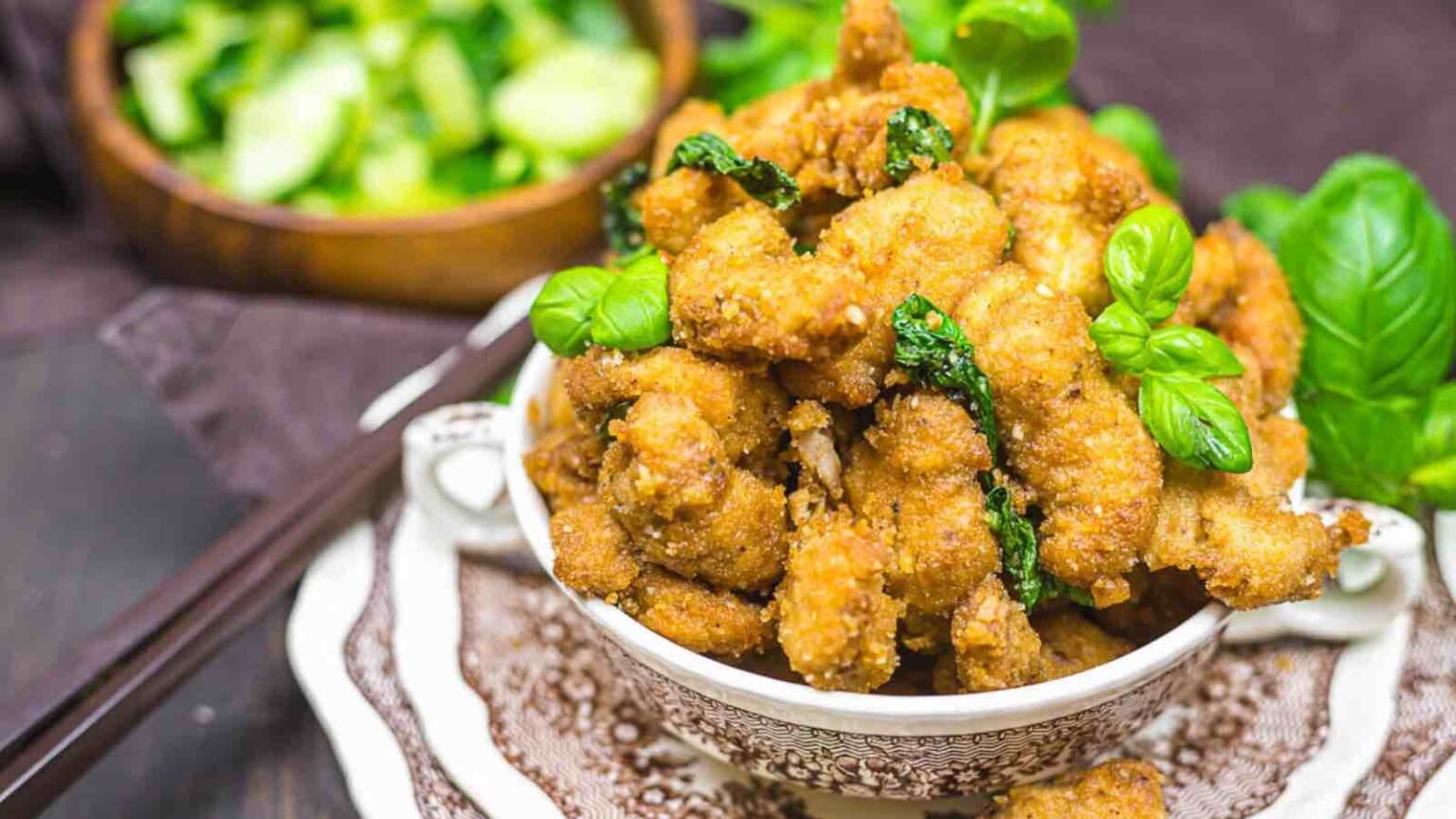 A bowl of golden-brown fried chicken pieces garnished with fresh basil leaves, with a bowl of sliced cucumbers in the background and chopsticks to the side.