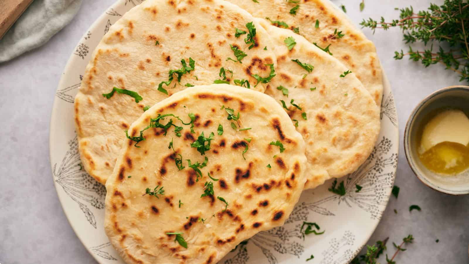 Three pieces of naan bread garnished with chopped herbs on a patterned plate, with a small bowl of melted butter on the side.