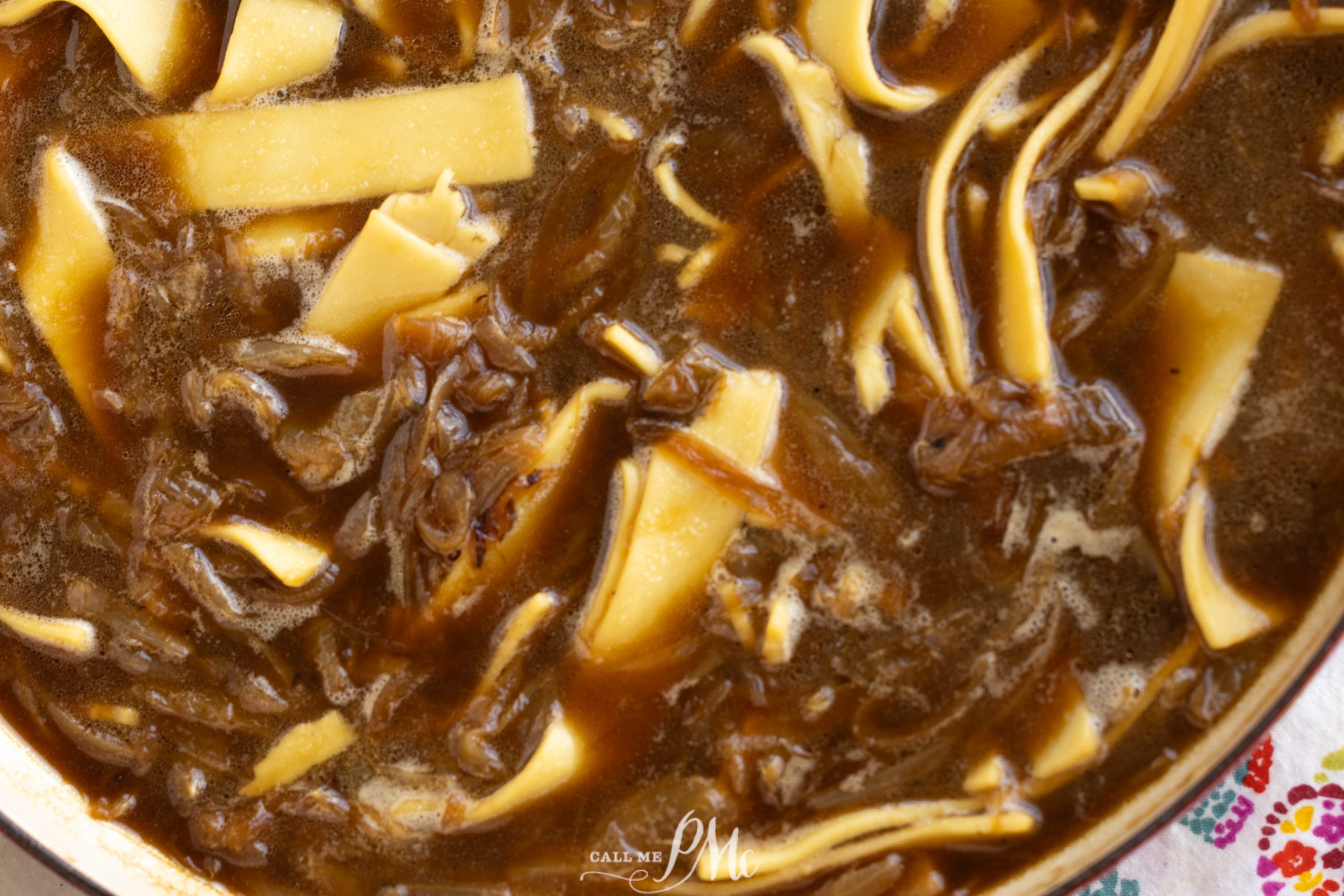 Close-up of beef and noodle soup with shredded beef and wide noodles in a dark brown broth.