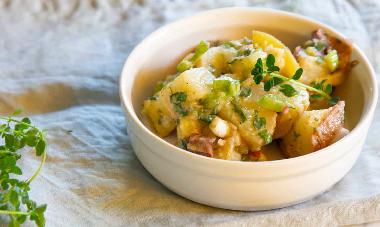White bowl filled with potato salad garnished with fresh herbs, placed on a light fabric surface with sprigs of herbs nearby.