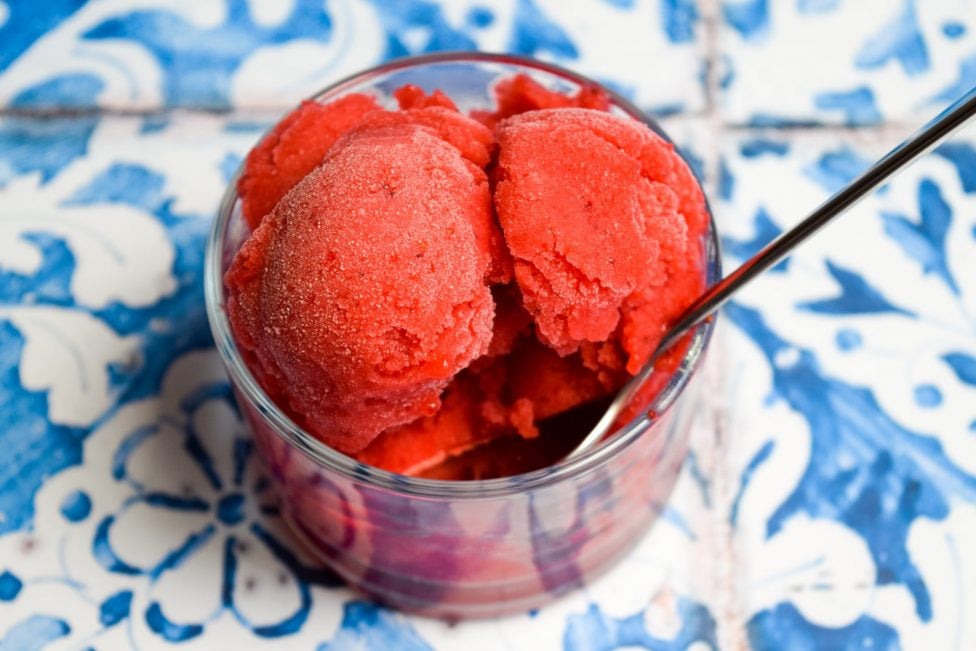 A glass bowl filled with scoops of bright red sorbet and a metal spoon, placed on a blue and white patterned surface.
