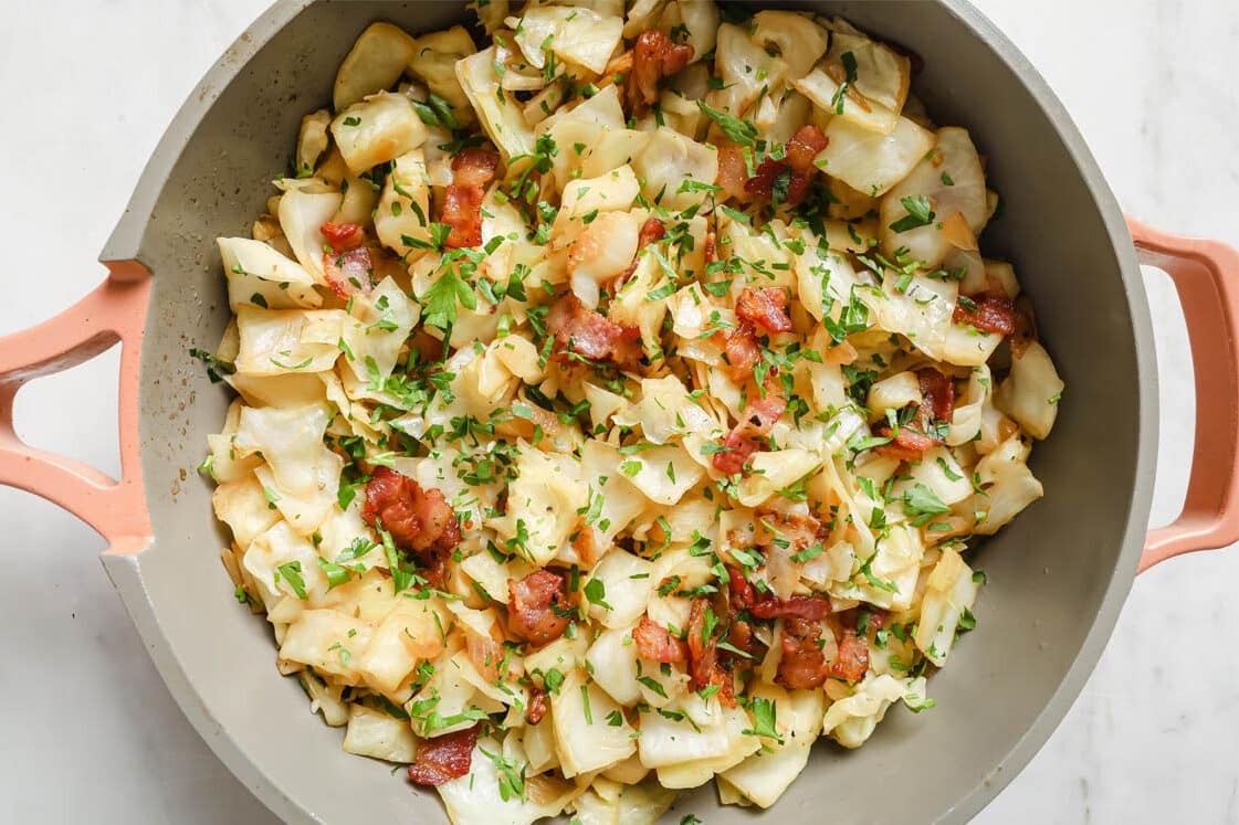 Sautéed cabbage with bacon pieces and chopped parsley in a large, light-colored skillet.