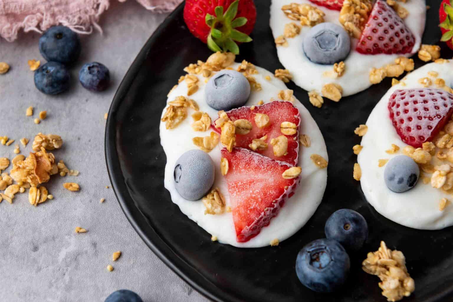 Yogurt clusters topped with sliced strawberries, blueberries, and granola are arranged on a black plate, with additional berries and granola scattered nearby.