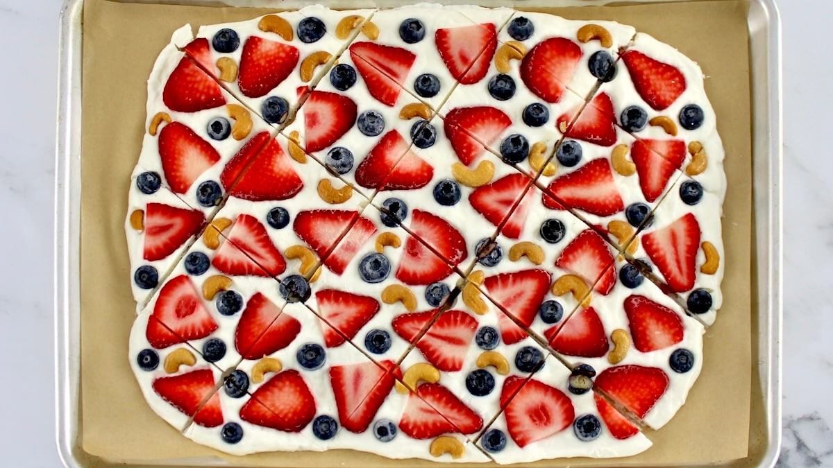 A rectangular sheet of dessert pizza topped with sliced strawberries, blueberries, and cashews, cut into square pieces on a baking tray.