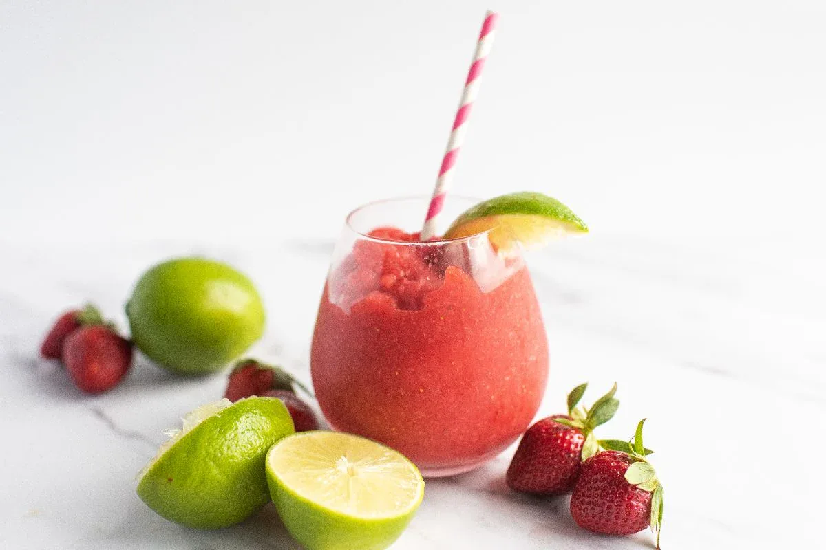 A glass of strawberry and lime smoothie with a striped straw, garnished with a lime wedge. Whole and halved limes and strawberries are placed around the glass on a white surface.