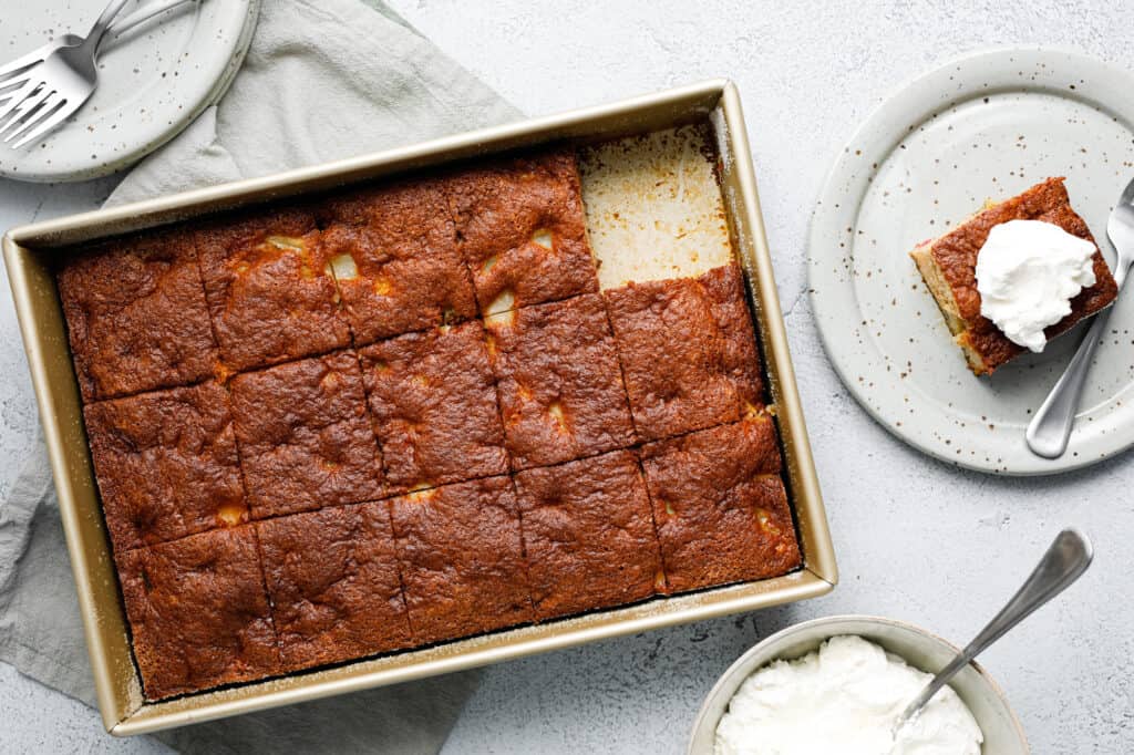 A rectangular pan of sliced cake, with one piece served on a plate topped with whipped cream; a bowl of whipped cream and forks are nearby.
