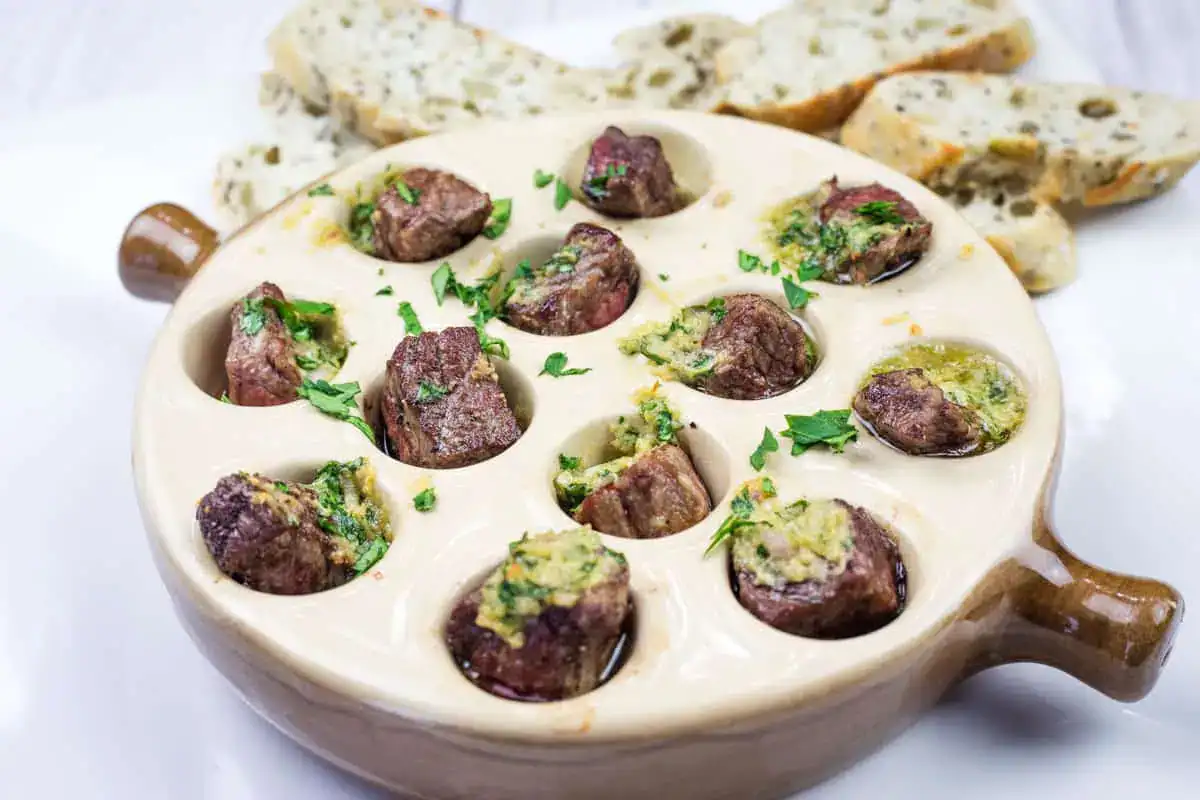 A ceramic escargot dish holds pieces of cooked meat topped with herbs and garlic butter, with sliced bread in the background.