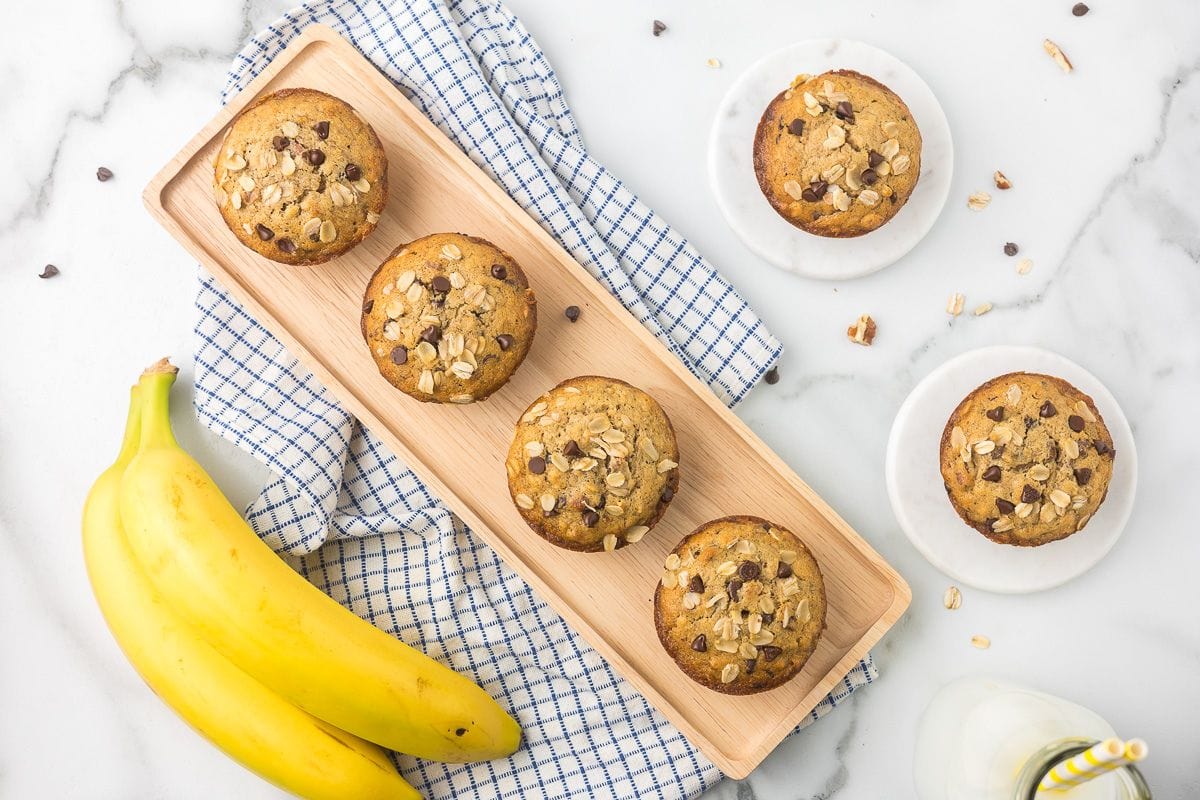 Five banana chocolate chip muffins on a wooden tray, two bananas, a glass of milk, and a checkered cloth on a white marble surface.