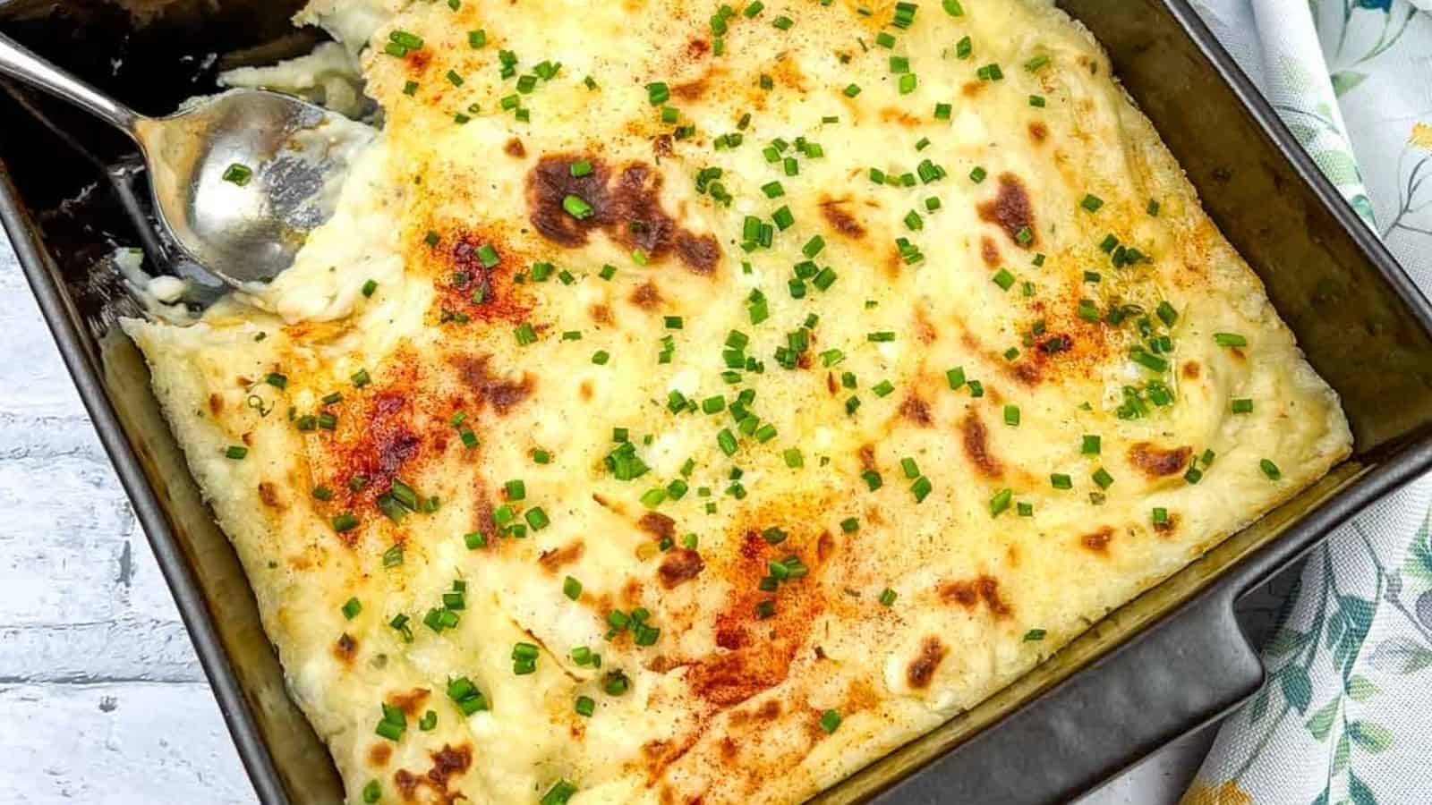 A baked casserole topped with browned mashed potatoes and chopped chives sits in a square baking dish with a serving spoon.