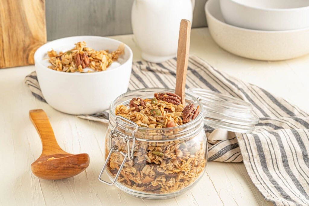 A glass jar filled with granola and pecans sits on a striped cloth next to a bowl of granola and a wooden spoon on a light-colored table.