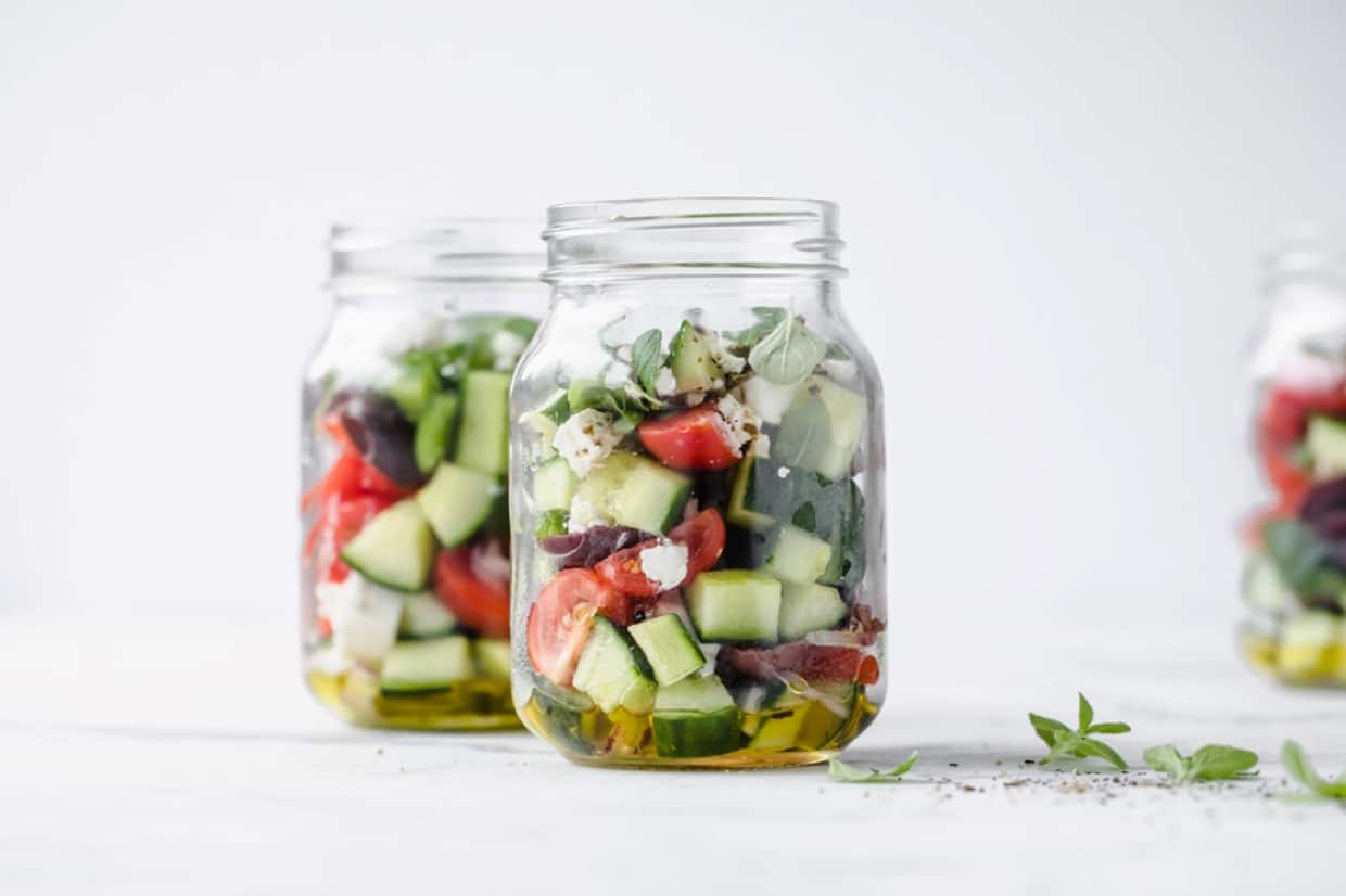 Three glass jars filled with a layered salad of chopped cucumbers, tomatoes, olives, feta cheese, herbs, and olive oil, placed on a white surface with a light background.