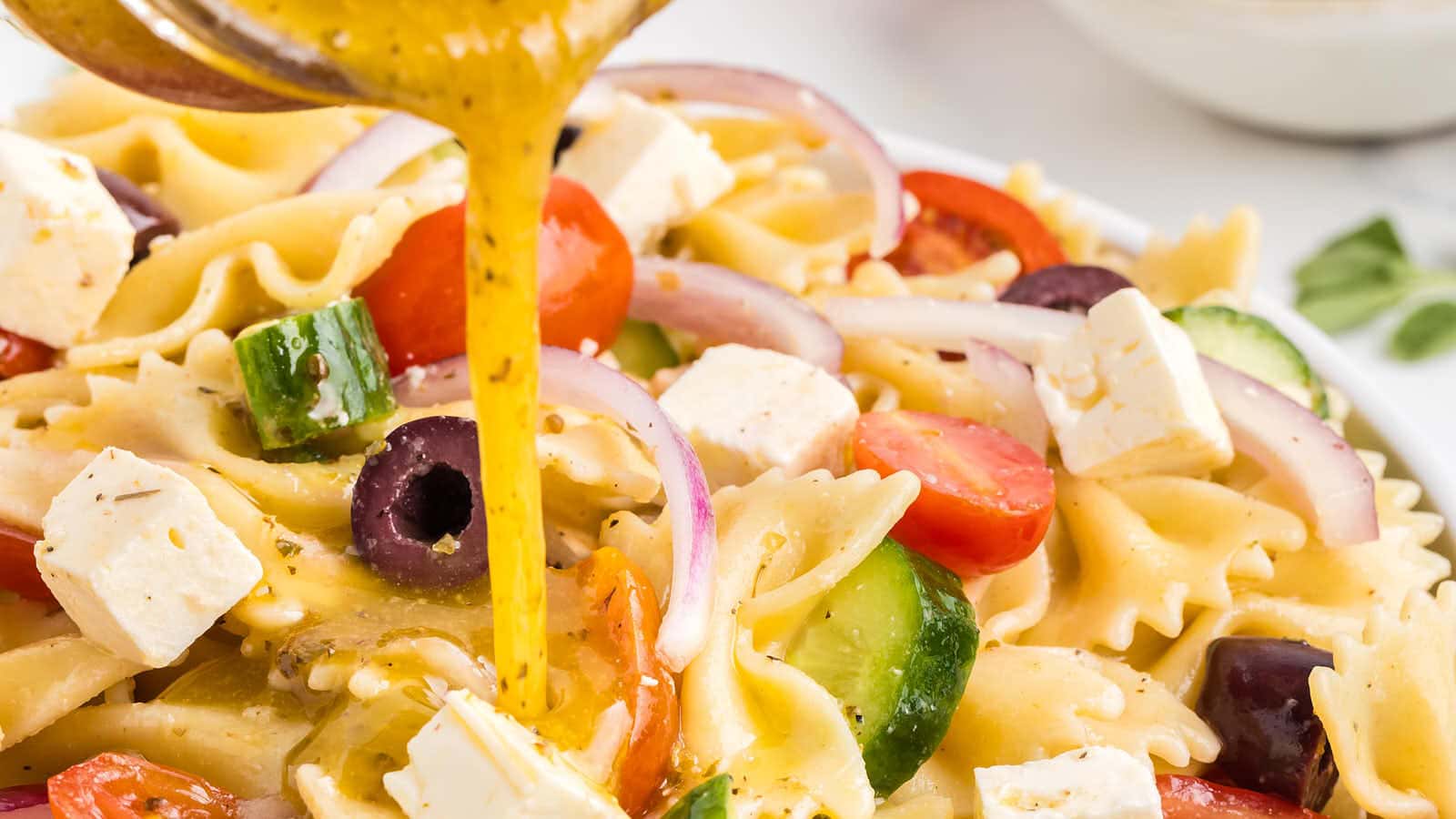 Close-up of Greek pasta salad with bowtie pasta, cherry tomatoes, cucumbers, red onion, olives, feta cheese, and vinaigrette being poured over the dish.