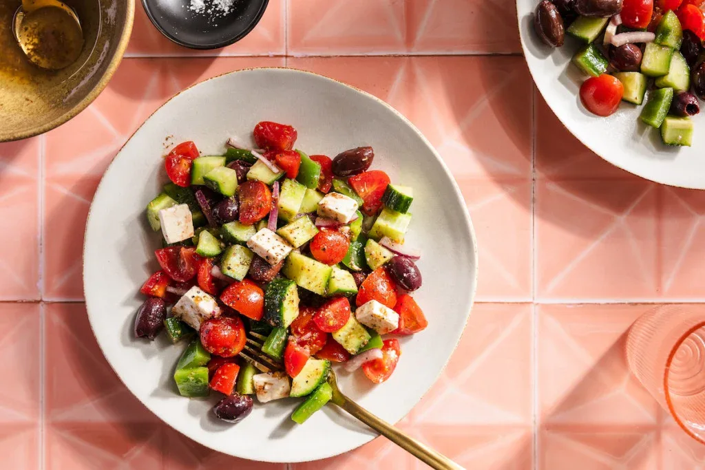 A plate of Greek salad with chopped cucumbers, tomatoes, feta cheese, and kalamata olives on a pink tiled surface; a fork rests on the plate.
