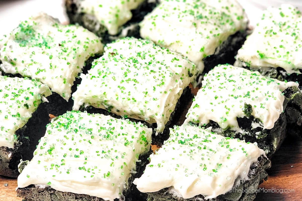 Nine square pieces of dark cake with white frosting and green sprinkles are arranged closely together on a wooden surface.
