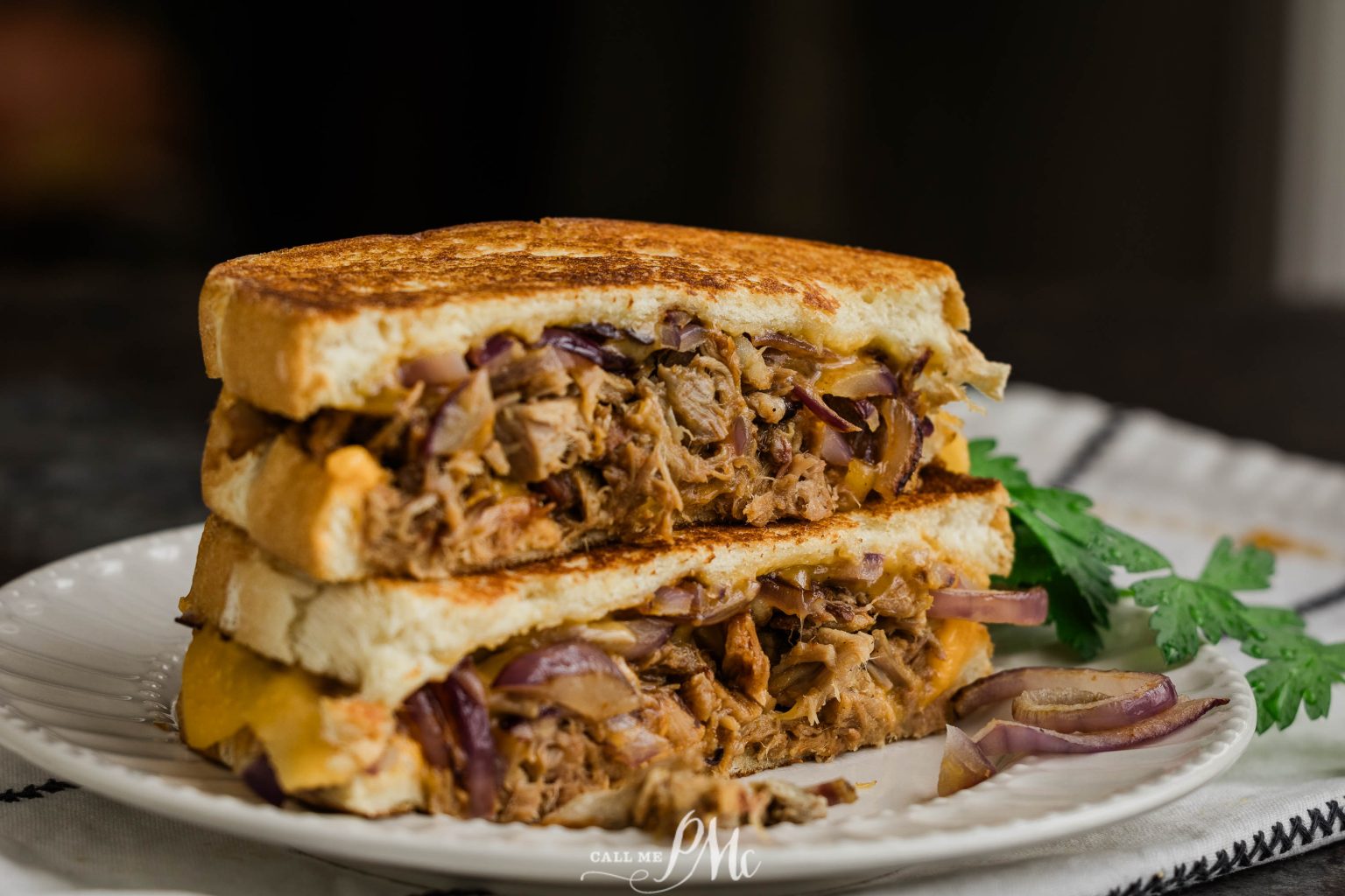 A close-up of a grilled sandwich cut in half, filled with shredded meat, melted cheese, and red onions, served on a white plate with a garnish on the side.
