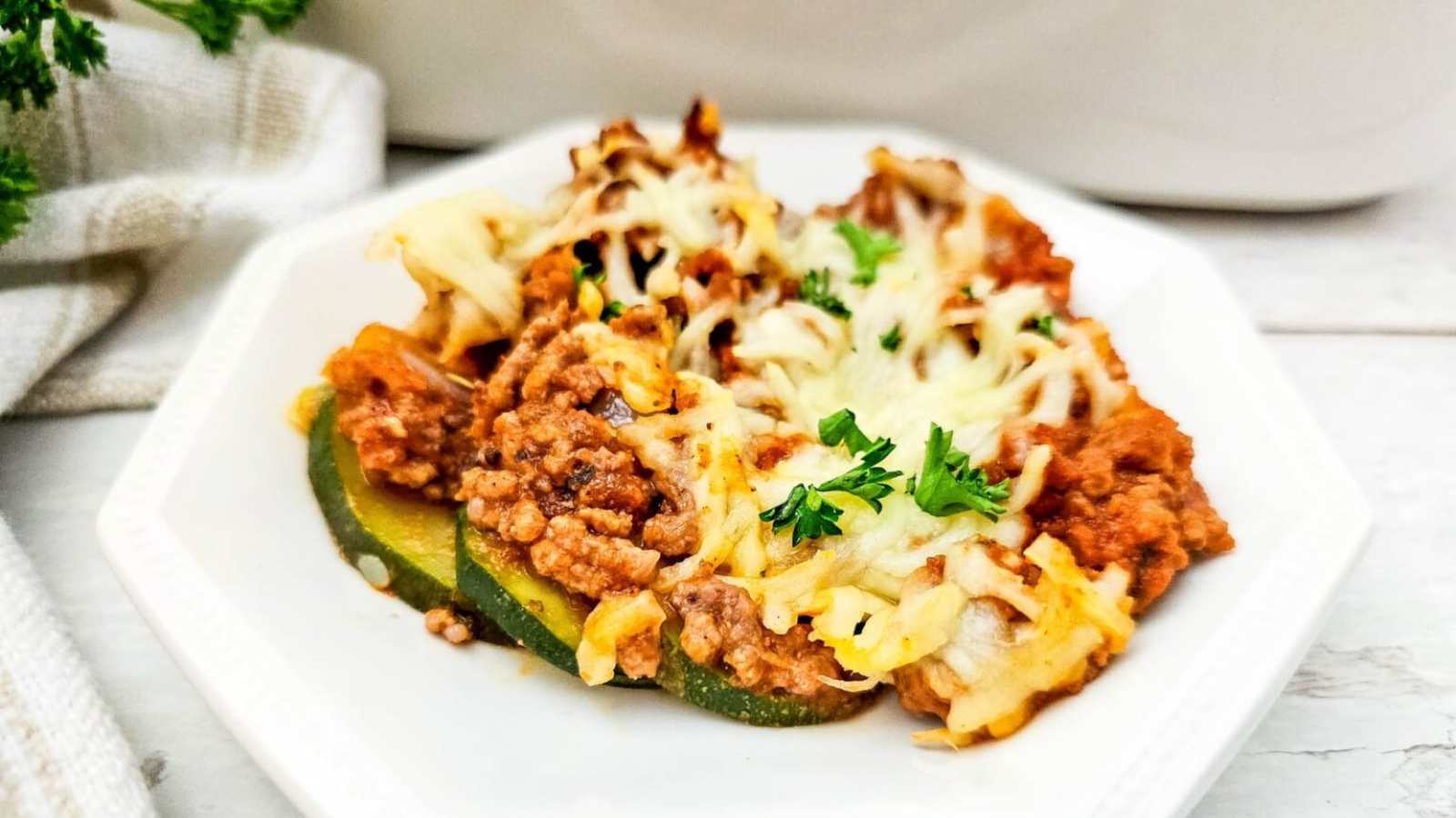 A serving of zucchini casserole topped with ground meat, tomato sauce, melted shredded cheese, and garnished with chopped parsley on a white plate.