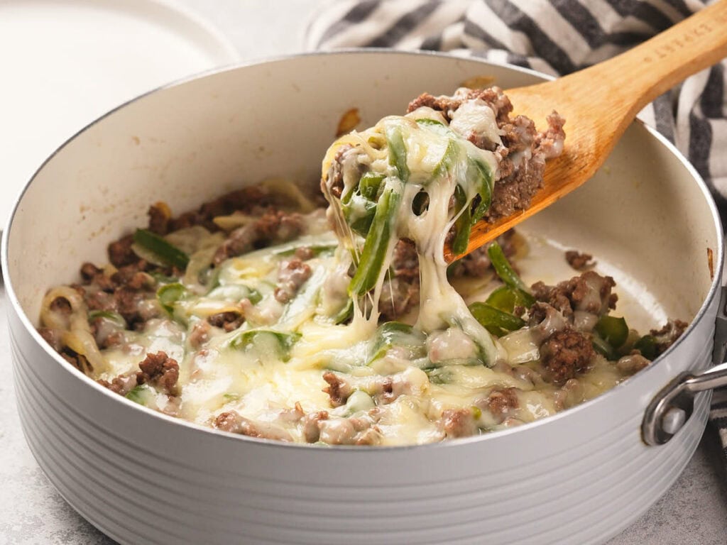 A skillet filled with ground beef, green peppers, and melted cheese is being stirred with a wooden spatula.