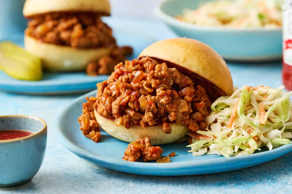 A sloppy joe sandwich with ground meat on a bun is served with coleslaw on a blue plate. A pickle spear, another sandwich, and sauce are visible in the background.