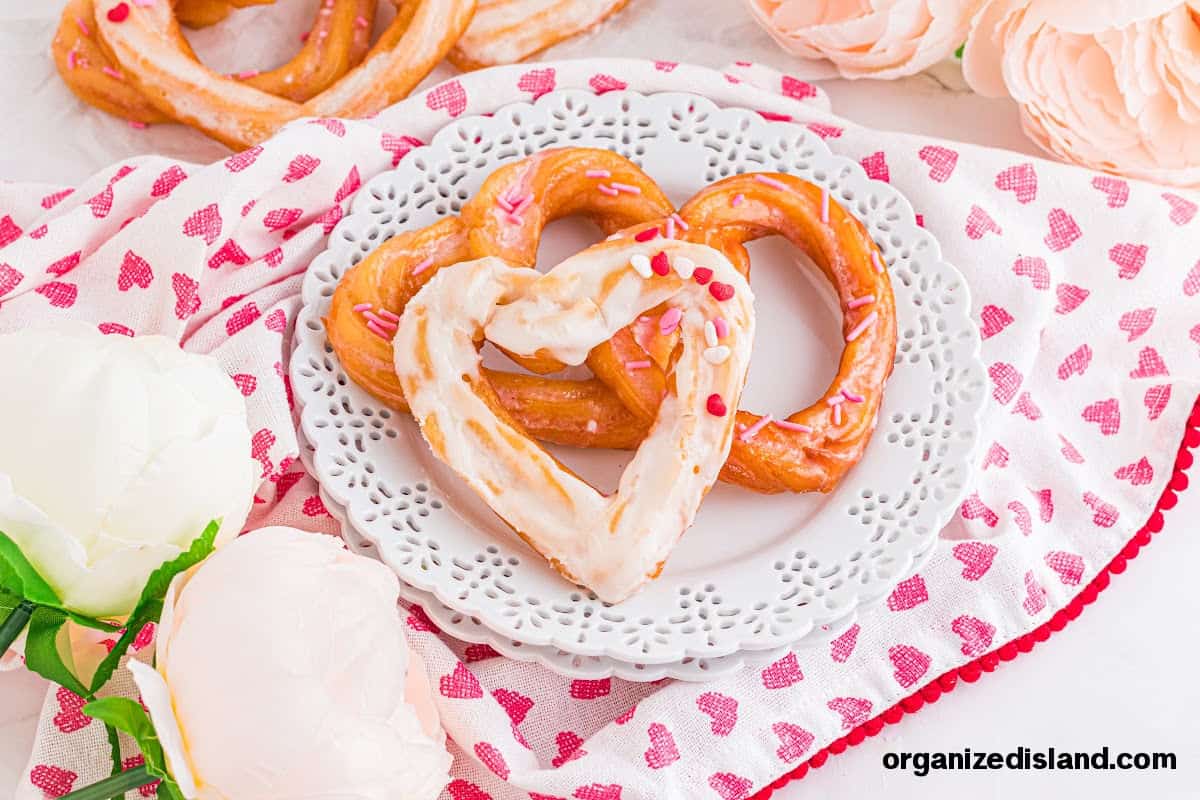 Heart-shaped pretzels with white icing and pink sprinkles on a decorative plate, surrounded by white flowers and a heart-patterned napkin.
