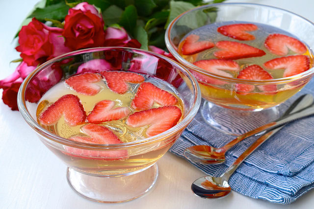 Two glass bowls filled with a clear yellow gelatin dessert topped with sliced strawberries, placed next to a bunch of red roses and two metal spoons on a blue napkin.