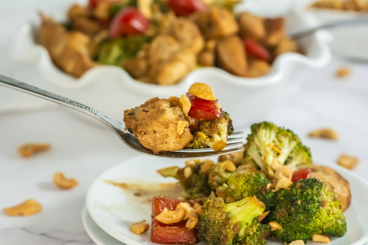 A fork holds a bite of stir-fried chicken, broccoli, and red bell pepper over a plate with more vegetables and nuts. A serving dish of the same food is in the background.