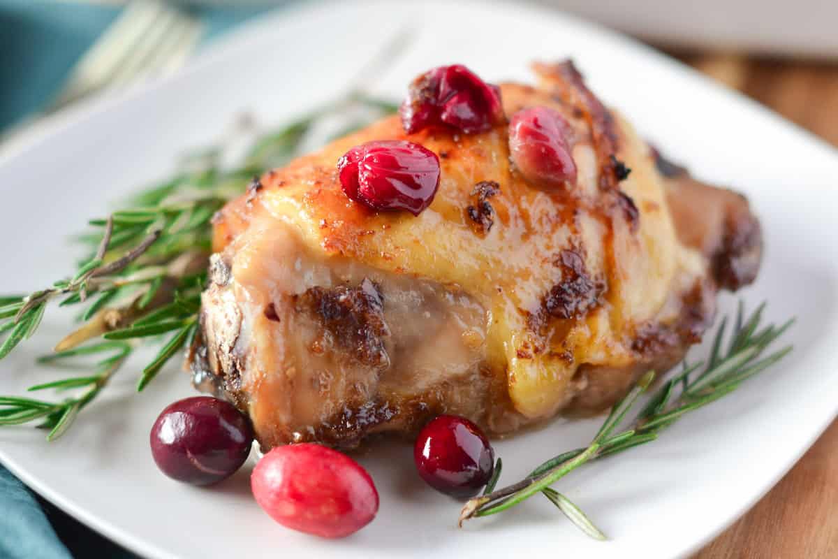 A roasted chicken thigh garnished with rosemary sprigs and cranberries on a white plate.