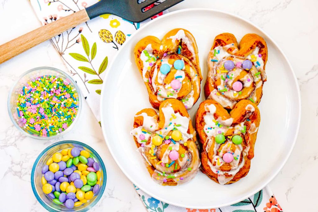 Four bunny-shaped cinnamon rolls with icing, pastel sprinkles, and colorful candies arranged on a white plate. Two bowls with additional sprinkles and candies are nearby.