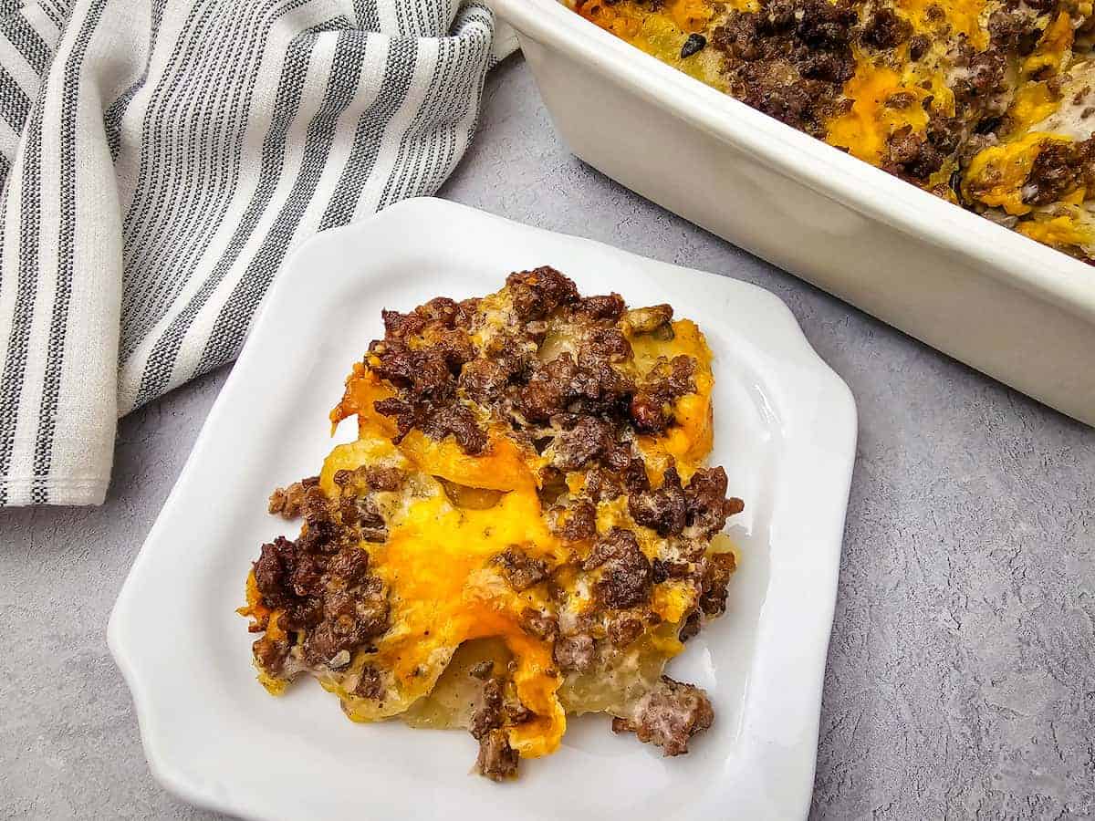 A serving of cheesy ground beef casserole on a white plate, with a striped towel and the casserole dish in the background.
